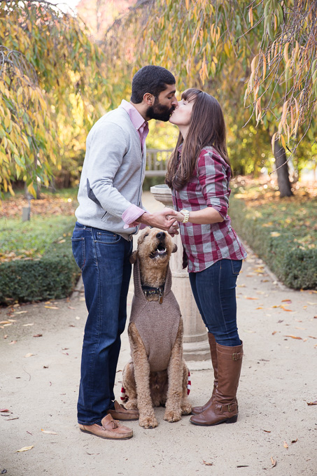husband and wife couple kissing in front of fountain with dog looking up in between them