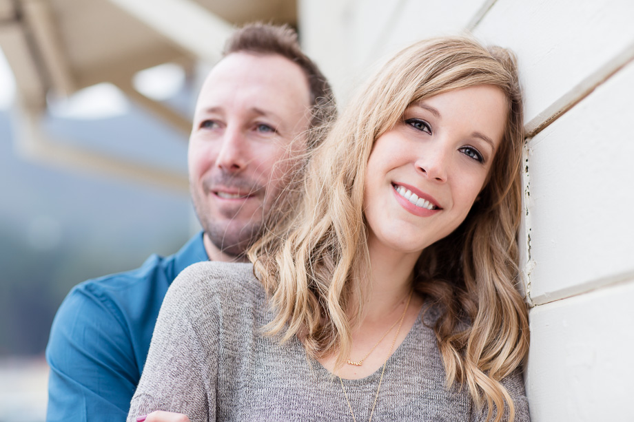 engagement photo of man behind his fiancee leaning on a building wall