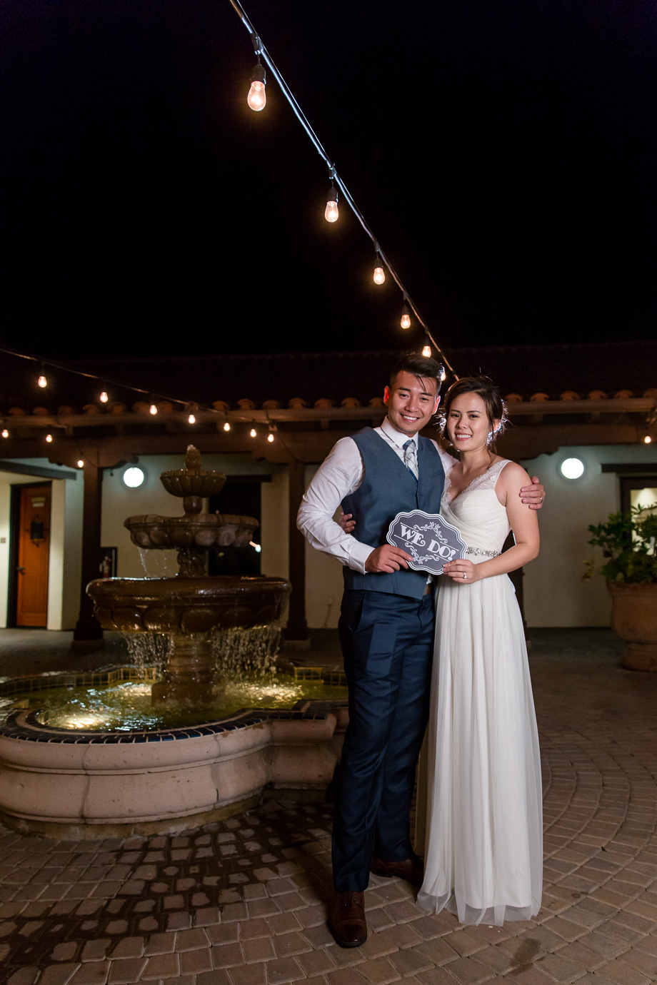 bride and groom holding the i do sign under the romantic cafe string lights